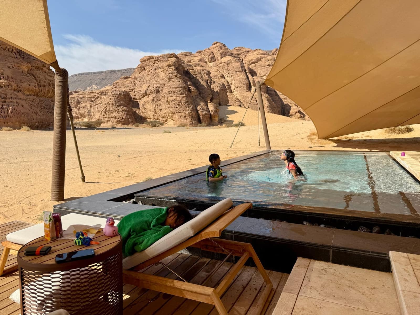 Sophie and George playing in a pool while Claire sleeps beside them in AlUla