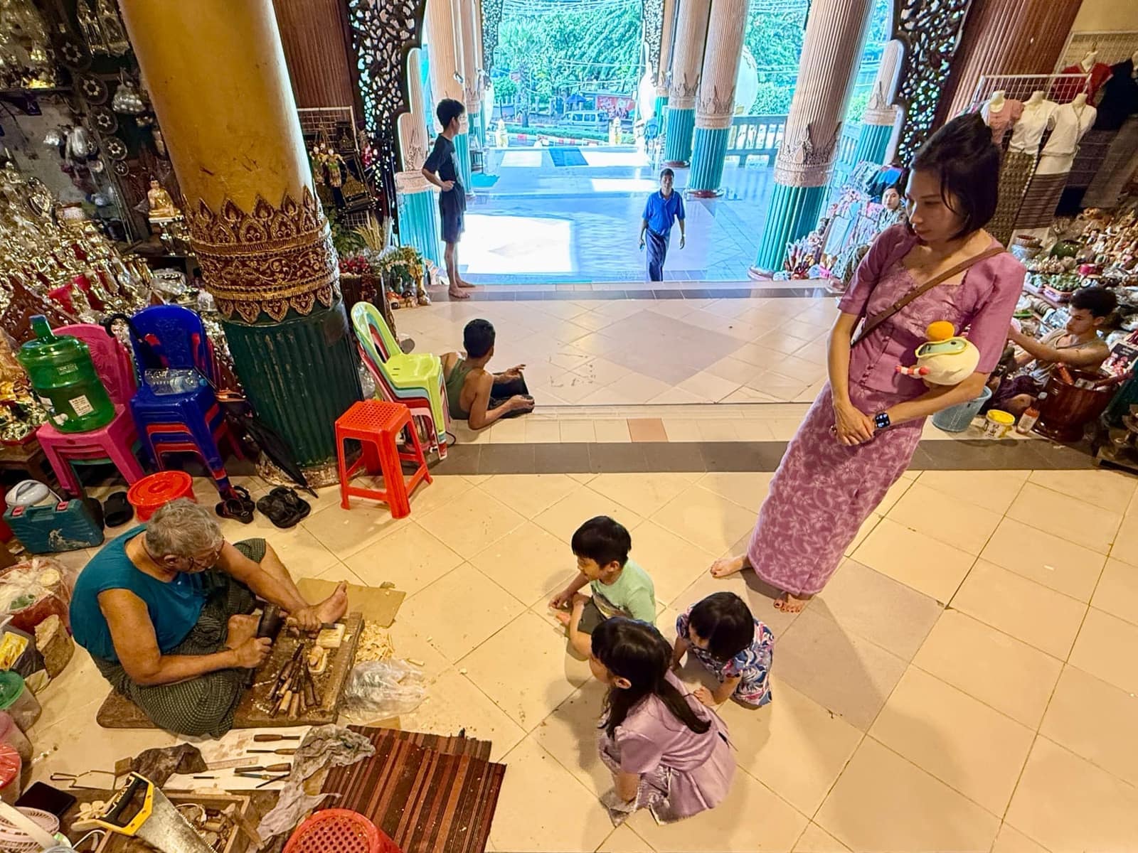 The three children seated beside a craftsman carving wooden figurines on the covered approach to Shwedagon Pagoda